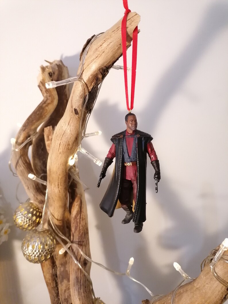 K&ouml;nnte beinhalten: Ein schwarzer und roter Star Wars-Charakter-Anh&auml;nger, der an einem Ast mit wei&szlig;en Lichtern h&auml;ngt. Der Charakter tr&auml;gt einen schwarzen Umhang und h&auml;lt einen Blaster.