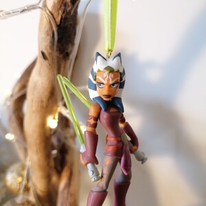 May include: A green lightsaber wielding Ahsoka Tano Star Wars Christmas ornament. The ornament is made of plastic and features a detailed sculpt of the character. The ornament is hanging from a green ribbon.