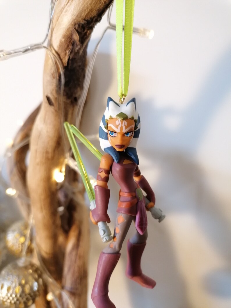 May include: A green lightsaber wielding Ahsoka Tano Star Wars Christmas ornament. The ornament is made of plastic and features a detailed sculpt of the character. The ornament is hanging from a green ribbon.