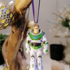 May include: A white and green plastic ornament of Buzz Lightyear from the Toy Story movie franchise. The ornament is hanging from a purple ribbon.