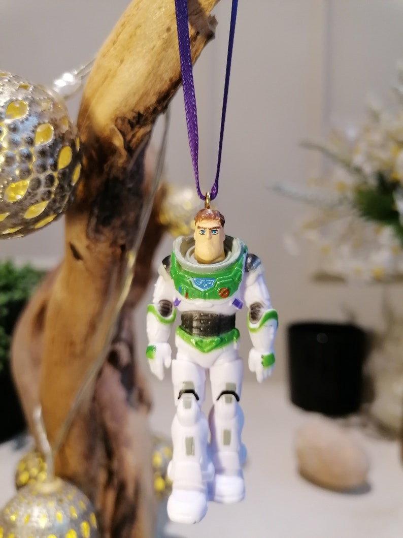 May include: A white and green plastic ornament of Buzz Lightyear from the Toy Story movie franchise. The ornament is hanging from a purple ribbon.