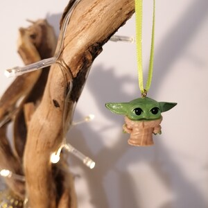 May include: A green and tan Baby Yoda ornament hanging from a light green ribbon. The ornament is suspended in front of a piece of driftwood decorated with small white lights. The ornament has large black eyes and a tan coat.