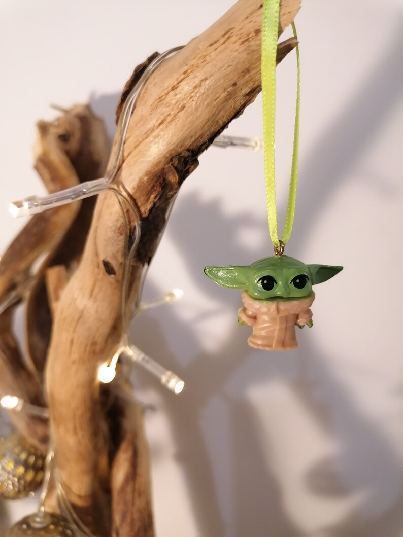 May include: A green and tan Baby Yoda ornament hanging from a light green ribbon. The ornament is suspended in front of a piece of driftwood decorated with small white lights. The ornament has large black eyes and a tan coat.