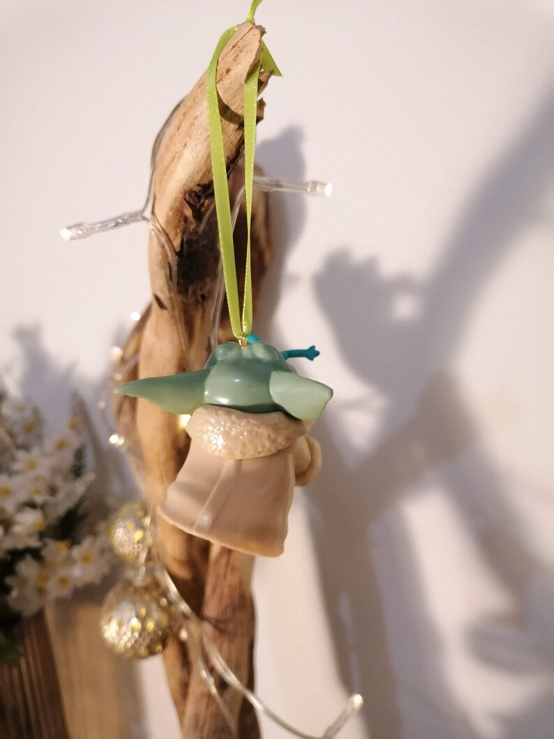 May include: A green and beige ornament of a popular character from the Star Wars franchise, The Mandalorian. The ornament is hanging from a green ribbon.