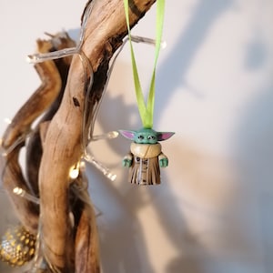Puede incluir: Una figurita verde y marr&oacute;n de un personaje de la franquicia Star Wars, colgando de una cinta verde en un trozo de madera a la deriva.