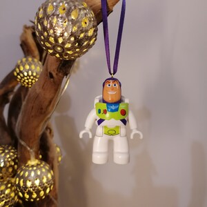 May include: A white plastic toy astronaut ornament with a purple, green, and yellow spacesuit, hanging from a purple ribbon. The astronaut has a smiling face and is wearing a helmet.
