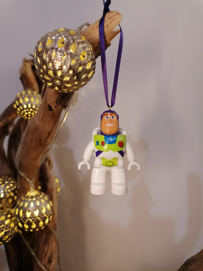 May include: A white plastic toy astronaut ornament with a purple, green, and yellow spacesuit, hanging from a purple ribbon. The astronaut has a smiling face and is wearing a helmet.