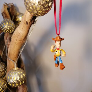 May include: A red ribbon hangs from a wooden branch with a plastic toy figurine of Woody from Toy Story. The figurine is wearing his signature cowboy hat, vest, and jeans. The branch is decorated with silver and gold string lights.