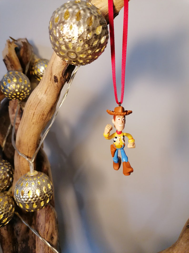 May include: A red ribbon hangs from a wooden branch with a plastic toy figurine of Woody from Toy Story. The figurine is wearing his signature cowboy hat, vest, and jeans. The branch is decorated with silver and gold string lights.