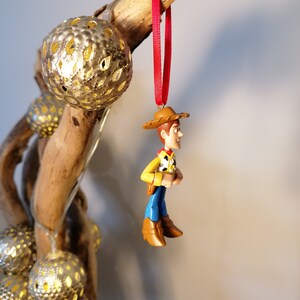 May include: A red and brown plastic ornament of Woody from Toy Story. Woody is wearing his signature cowboy hat, vest, and jeans. He is hanging from a red ribbon.