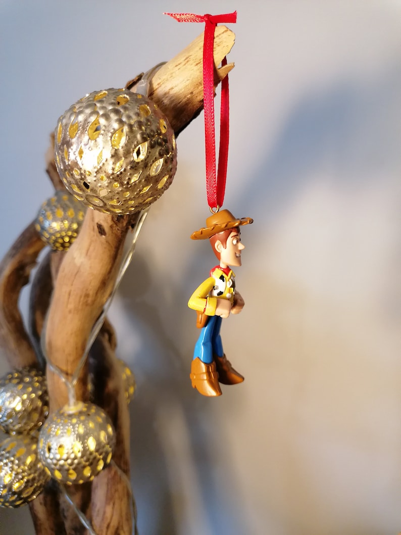 May include: A red and brown plastic ornament of Woody from Toy Story. Woody is wearing his signature cowboy hat, vest, and jeans. He is hanging from a red ribbon.