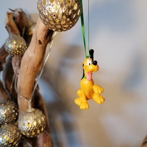 Op de afbeelding: Een gele en bruine cartoonhond ornament hangend aan een groen lint. Het ornament is een personage uit een populaire animatiefilm.