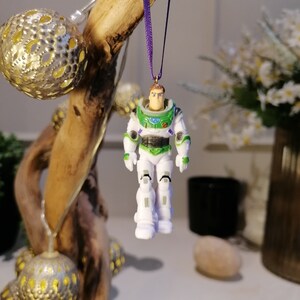 May include: A white and green plastic toy astronaut ornament with a purple ribbon hanging from a wooden branch. The astronaut has a black visor and a green space suit.
