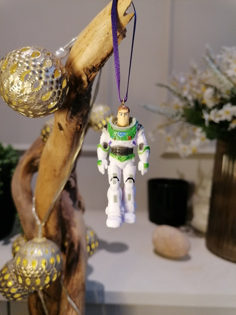 May include: A white and green plastic toy astronaut ornament with a purple ribbon hanging from a wooden branch. The astronaut has a black visor and a green space suit.