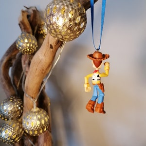 May include: A blue and brown plastic ornament of Woody from Toy Story. The ornament is hanging from a blue ribbon on a branch with silver and gold decorative lights.