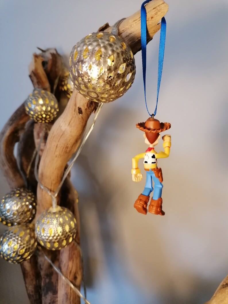 May include: A blue and brown plastic ornament of Woody from Toy Story. The ornament is hanging from a blue ribbon on a branch with silver and gold decorative lights.