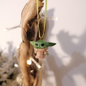 May include: A green and tan figurine of a character from a popular science fiction series, hanging from a light green ribbon. The figurine is suspended in front of a piece of driftwood, with a string of small white lights wrapped around it.