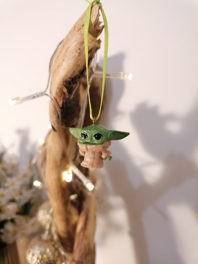 May include: A green and tan figurine of a character from a popular science fiction series, hanging from a light green ribbon. The figurine is suspended in front of a piece of driftwood, with a string of small white lights wrapped around it.