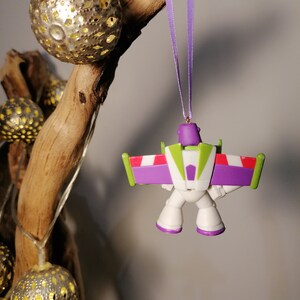 May include: A purple and white plastic ornament of Buzz Lightyear from the Toy Story franchise. The ornament is hanging from a purple ribbon and is in a flying pose.