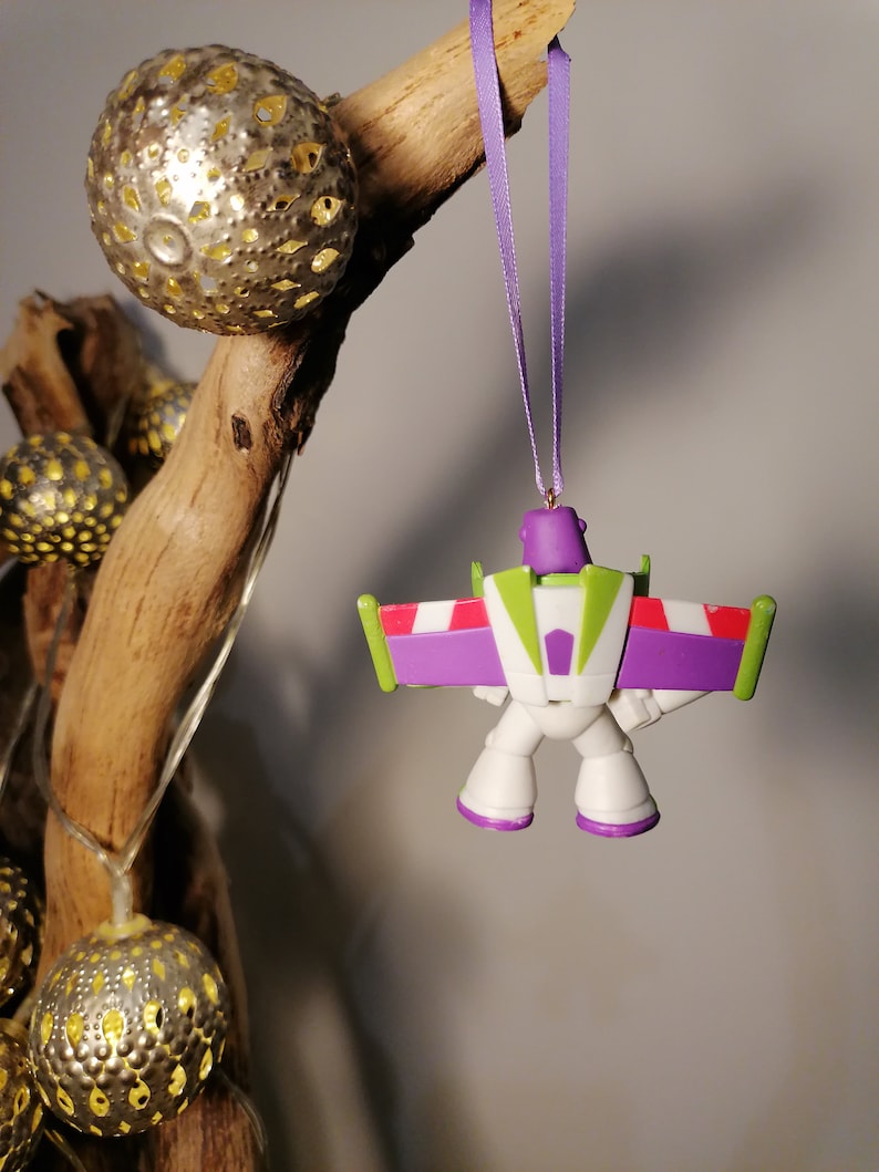 May include: A purple and white plastic ornament of Buzz Lightyear from the Toy Story franchise. The ornament is hanging from a purple ribbon and is in a flying pose.