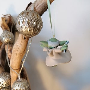 May include: A green and beige Yoda ornament hanging from a branch with a green ribbon. The ornament is holding a lightsaber in one hand.