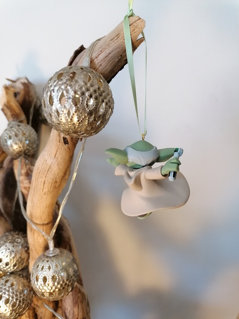May include: A green and beige Yoda ornament hanging from a branch with a green ribbon. The ornament is holding a lightsaber in one hand.