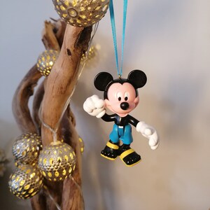 May include: A blue and yellow Mickey Mouse Christmas ornament hanging from a branch with string lights. The ornament is made of plastic and has a 3D design.