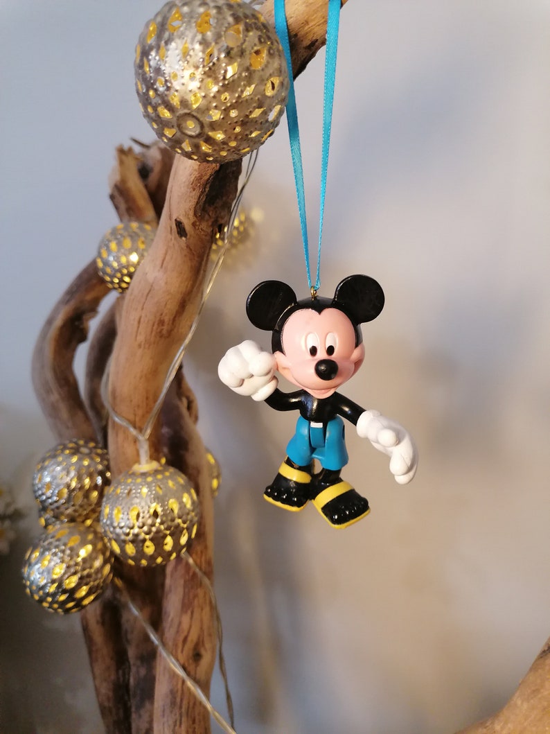 May include: A blue and yellow Mickey Mouse Christmas ornament hanging from a branch with string lights. The ornament is made of plastic and has a 3D design.