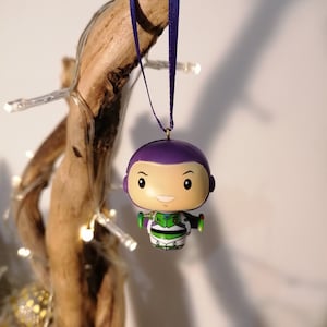 Puede incluir: Adorno navideño morado y verde con un personaje de dibujos animados con un casco morado y un traje espacial verde y blanco. El adorno está colgado de una cinta morada.