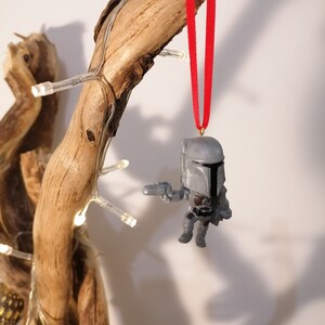 May include: A grey and black Star Wars character ornament hanging from a red ribbon on a branch. The ornament is a small, detailed figure of a Mandalorian.
