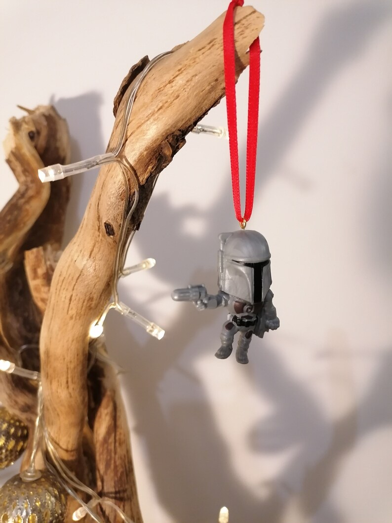 May include: A grey and black Star Wars character ornament hanging from a red ribbon on a branch. The ornament is a small, detailed figure of a Mandalorian.