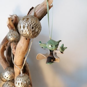 May include: A green Yoda character ornament hanging from a branch with a green ribbon. The ornament is made of plastic and has a detailed design. The branch is decorated with silver and white ornaments.