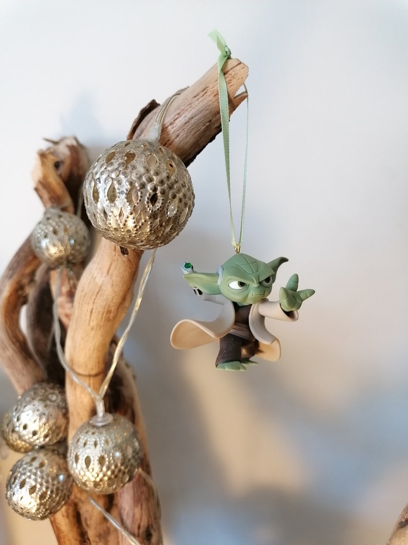 May include: A green Yoda character ornament hanging from a branch with a green ribbon. The ornament is made of plastic and has a detailed design. The branch is decorated with silver and white ornaments.