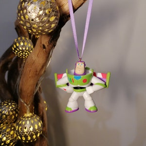 May include: A purple ribbon hangs from a branch with a plastic Buzz Lightyear ornament attached. The ornament is green, white, and red with a black belt and a purple face.