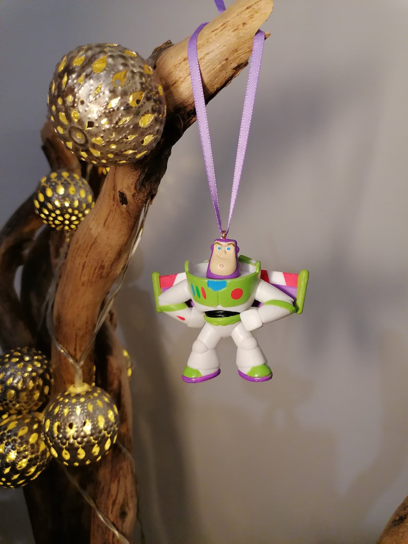 May include: A purple ribbon hangs from a branch with a plastic Buzz Lightyear ornament attached. The ornament is green, white, and red with a black belt and a purple face.
