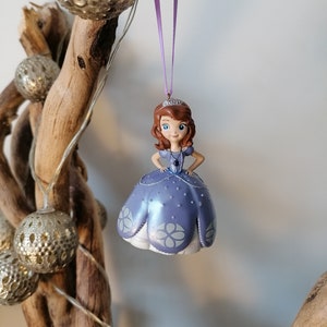 May include: A purple and white princess ornament with a crown, brown hair, and blue eyes. The ornament is hanging from a purple ribbon.