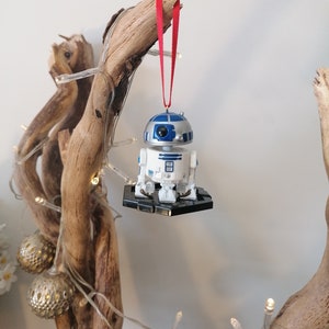 May include: A white and blue R2-D2 Star Wars character ornament hanging from a branch with a red ribbon. The ornament is on a black base and has a silver finish.