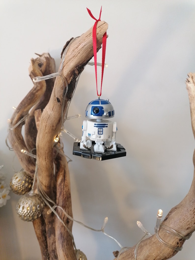 May include: A white and blue R2-D2 Star Wars character ornament hanging from a branch with a red ribbon. The ornament is on a black base and has a silver finish.