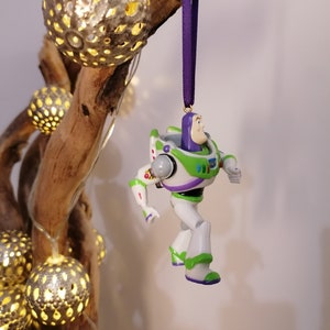 May include: A green and white plastic toy figurine of Buzz Lightyear from the Toy Story franchise. The figurine is hanging from a purple ribbon and is posed with one leg raised and his arms outstretched.