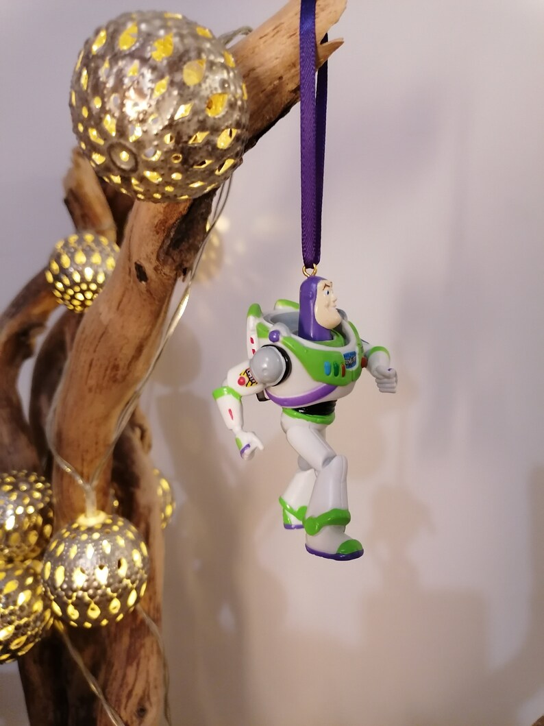 May include: A green and white plastic toy figurine of Buzz Lightyear from the Toy Story franchise. The figurine is hanging from a purple ribbon and is posed with one leg raised and his arms outstretched.