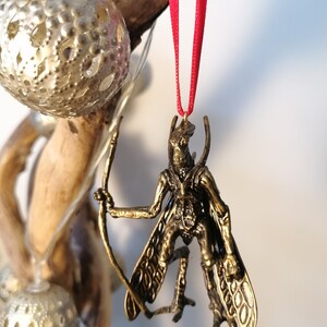 May include: A gold-coloured ornament depicting a winged creature with a bow and arrow. The ornament is hanging from a red ribbon.