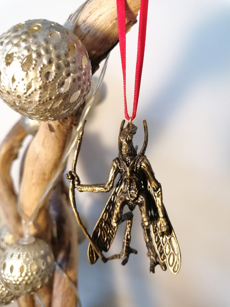 May include: A gold-coloured ornament depicting a winged creature with a bow and arrow. The ornament is hanging from a red ribbon.