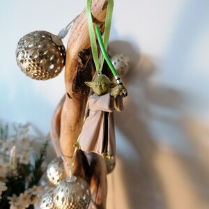 Puede incluir: Adorno navide&ntilde;o de Yoda con un sable de luz verde y una cinta verde para colgar. La figura de Yoda lleva una t&uacute;nica marr&oacute;n. El adorno est&aacute; colgado de un trozo de madera flotante, con luces decorativas plateadas.