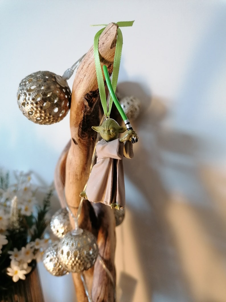 Puede incluir: Adorno navide&ntilde;o de Yoda con un sable de luz verde y una cinta verde para colgar. La figura de Yoda lleva una t&uacute;nica marr&oacute;n. El adorno est&aacute; colgado de un trozo de madera flotante, con luces decorativas plateadas.