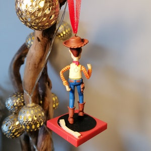 Puede incluir: Un adorno navide&ntilde;o rojo y blanco con una figura de pl&aacute;stico de Woody de Toy Story. Woody lleva su caracter&iacute;stico sombrero de vaquero, chaleco y pantalones vaqueros. Est&aacute; de pie sobre una base circular roja y negra.