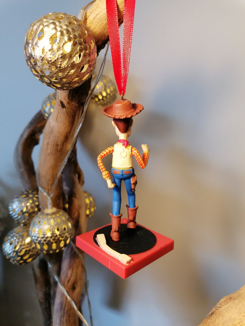 Puede incluir: Un adorno navide&ntilde;o rojo y blanco con una figura de pl&aacute;stico de Woody de Toy Story. Woody lleva su caracter&iacute;stico sombrero de vaquero, chaleco y pantalones vaqueros. Est&aacute; de pie sobre una base circular roja y negra.