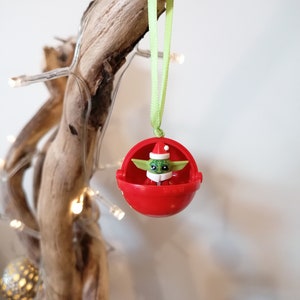 Pu&ograve; includere: Un addobbo natalizio rosso con una piccola figura verde che indossa un cappello da Babbo Natale all'interno. L'addobbo &egrave; appeso a un nastro verde su un ramo.