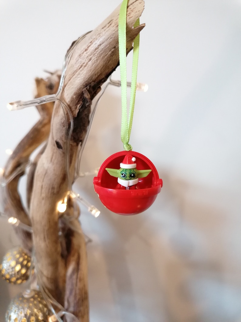 Pu&ograve; includere: Un addobbo natalizio rosso con una piccola figura verde che indossa un cappello da Babbo Natale all'interno. L'addobbo &egrave; appeso a un nastro verde su un ramo.
