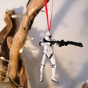 Puede incluir: Un adorno de pl&aacute;stico blanco de un Stormtrooper de Star Wars colgado de una rama con una cinta roja. La figura sostiene un rifle bl&aacute;ster negro.