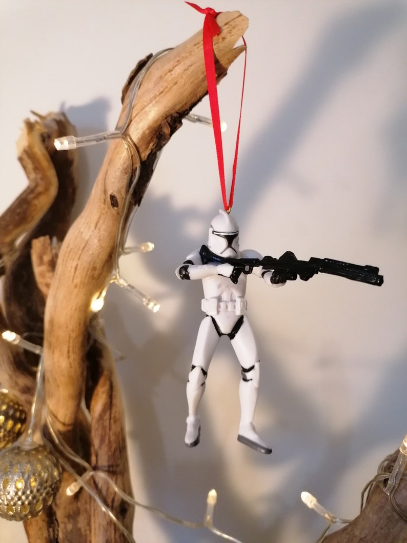 Puede incluir: Un adorno de pl&aacute;stico blanco de un Stormtrooper de Star Wars colgado de una rama con una cinta roja. La figura sostiene un rifle bl&aacute;ster negro.